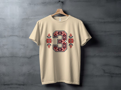 Beige t-shirt hanging on a wooden hanger against a textured gray wall, featuring a decorative red and blue number three graphic with cross-stitch patterns.