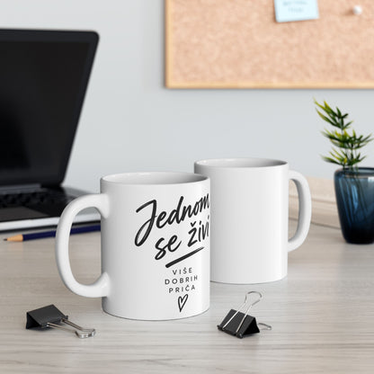 White mug with black text that says "Jednom se živi" and "Više dobrih priča" with a heart.