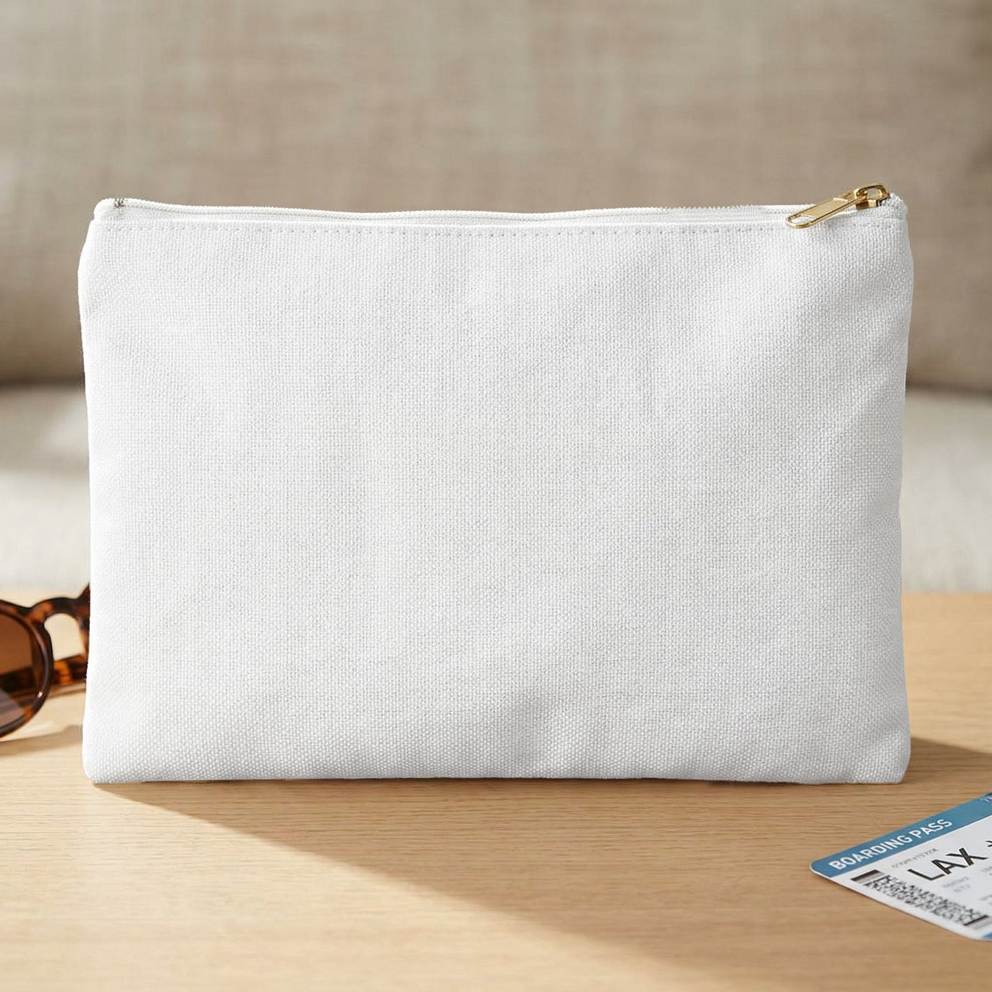 White canvas toiletry bag with gold zipper, sunglasses, and boarding pass on table.