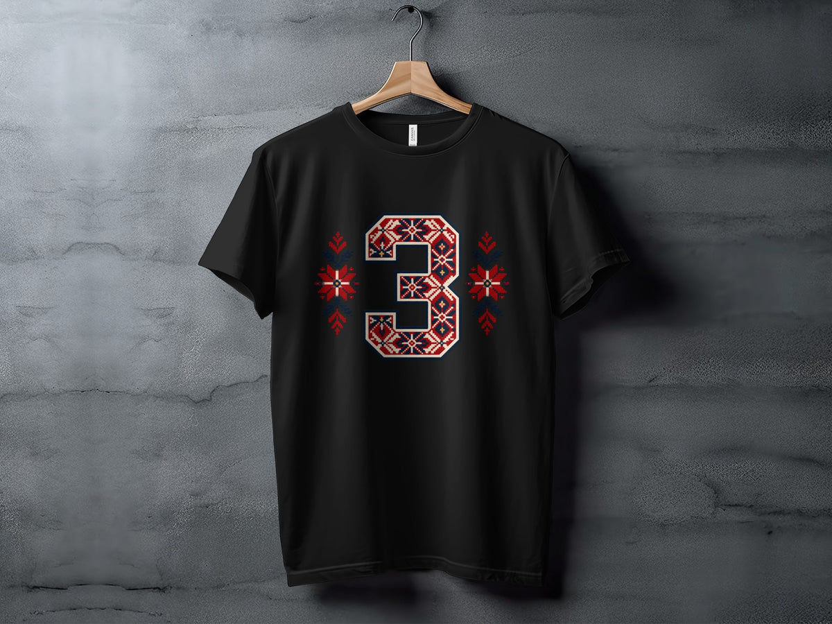 A black crewneck t-shirt hangs on a wooden hanger against a textured concrete wall, featuring a large number three in a red, white, and blue Nordic knit pattern on the chest, flanked by matching snowflake motifs.