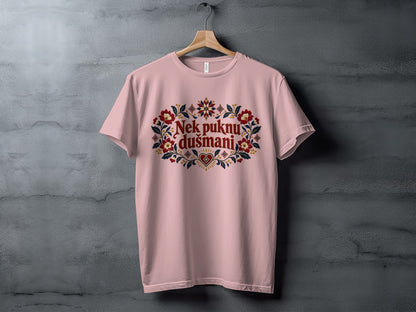 A pink crewneck t-shirt hangs on a wooden hanger against a textured concrete wall, featuring floral embroidery framing the text Nek puknu dušmani.
