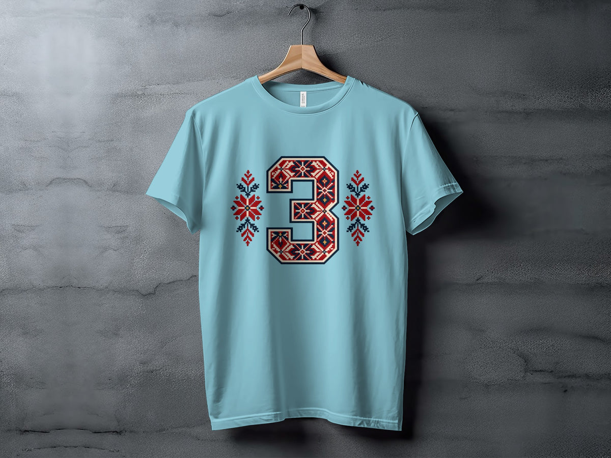 Light blue crewneck t-shirt hanging on a wooden hanger against a gray textured wall, featuring a large number three with red and blue Nordic embroidery.