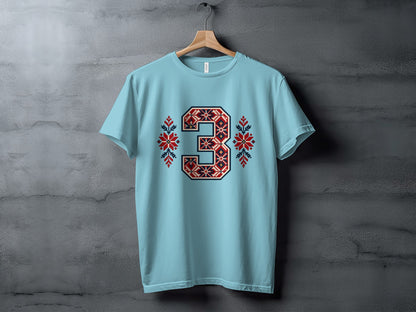 Light blue crewneck t-shirt hanging on a wooden hanger against a gray textured wall, featuring a large number three with red and blue Nordic embroidery.