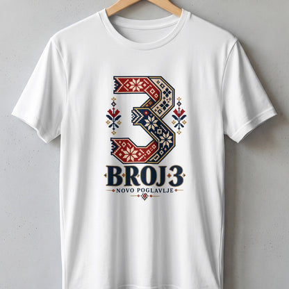 A white crewneck t-shirt hanging on a wooden hanger against a grey concrete wall, featuring a large number three printed in red and blue traditional folklore patterns with the text BROJ 3 NOVO POGLAVLJE underneath.