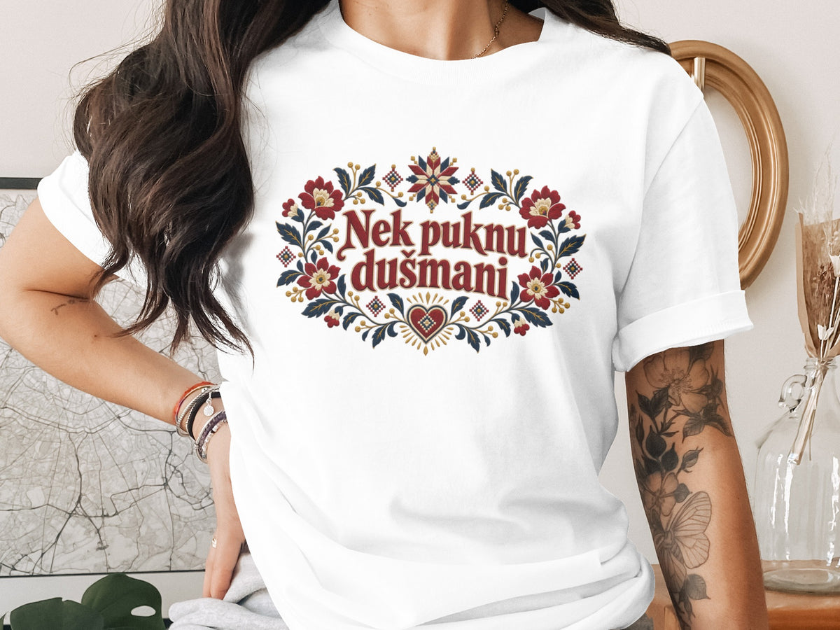 A person wearing a white t-shirt printed with red text reading Nek puknu dušmani surrounded by folkloric floral embroidery.