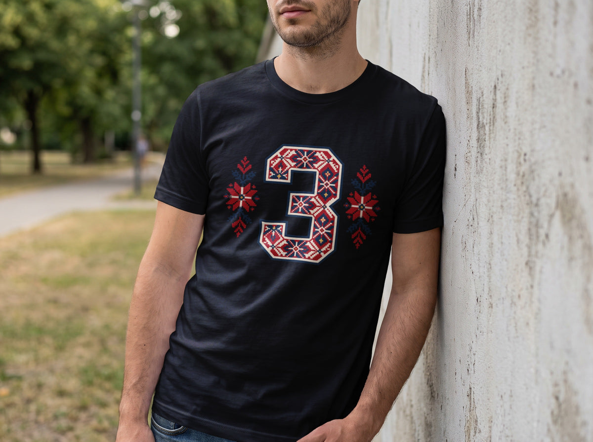 A man leaning against a concrete wall wearing a black t-shirt with a patterned number three.