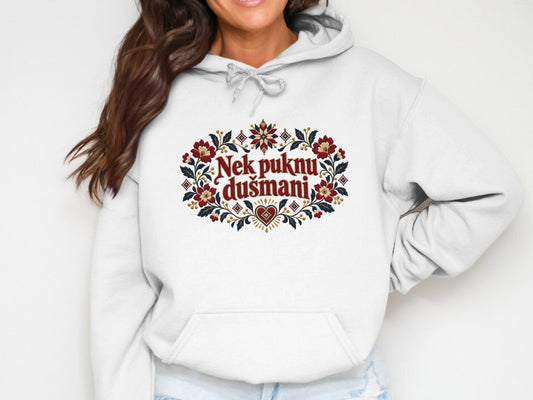 A person wearing a white hooded sweatshirt featuring embroidered red and blue floral motifs surrounding the text Nek puknu dušmani.