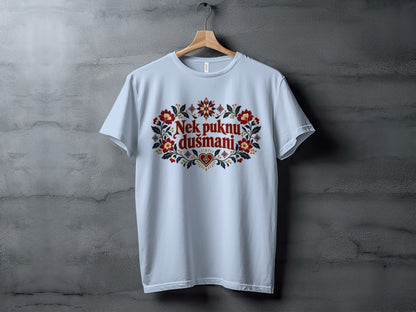 Light blue t-shirt hanging on a wooden hanger against a textured gray wall, featuring red floral embroidery and text reading Nek puknu dušmani.