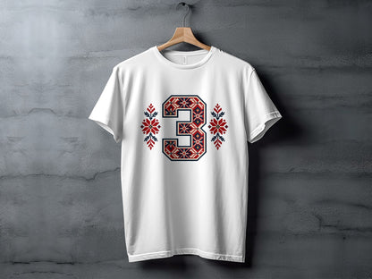 A white crewneck t-shirt hangs on a wooden hanger against a gray textured background, featuring a large number 3 in a red, blue, and white geometric pattern on the chest, flanked by matching snowflake designs.