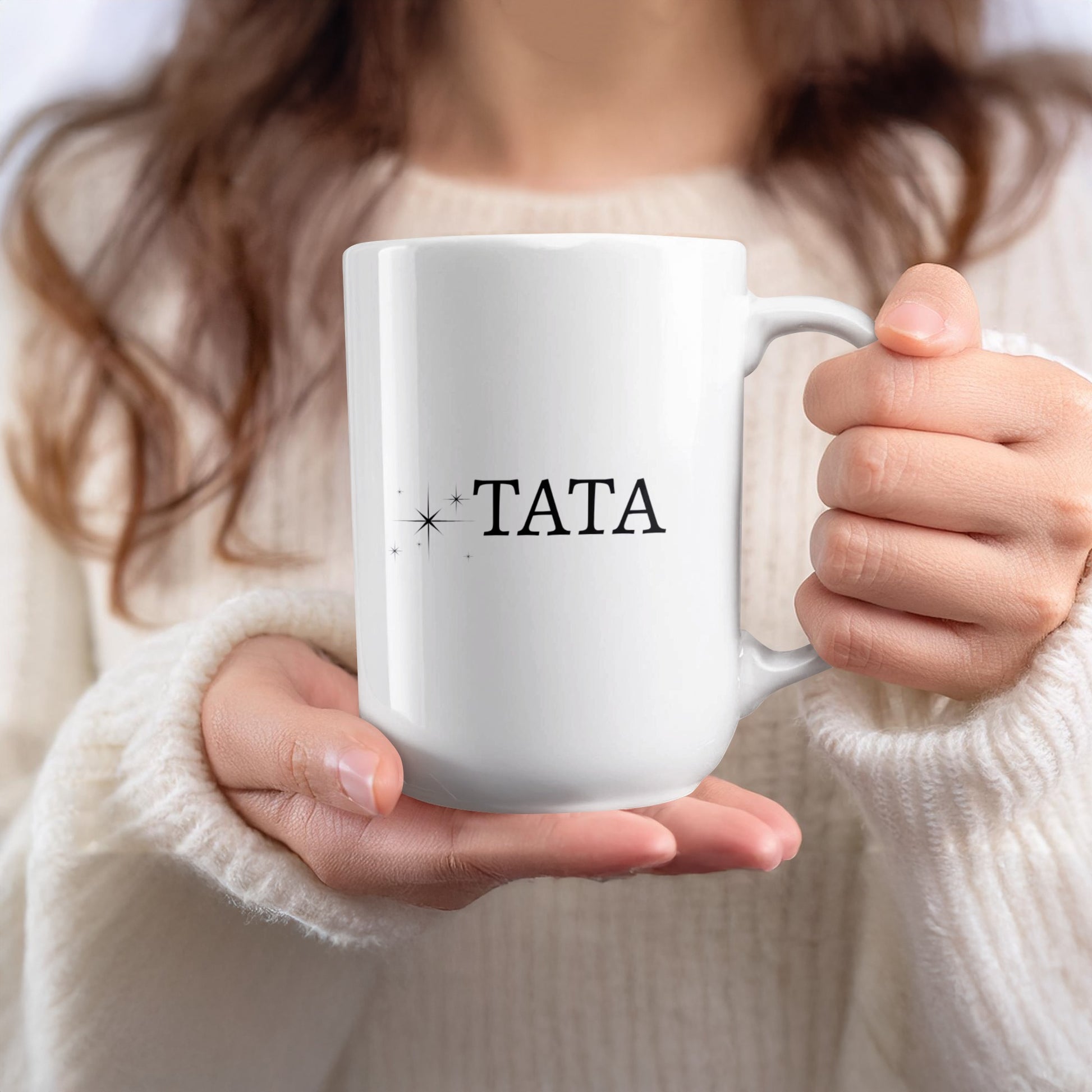 White mug with "TATA" and star accents held by a person in a cream sweater.