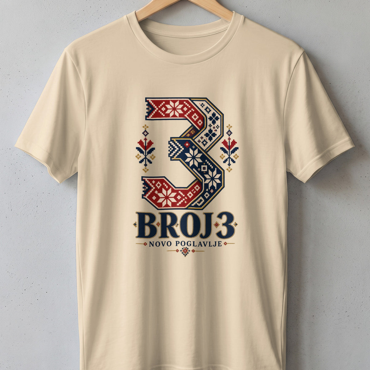Cream-colored crewneck t-shirt hanging on a wooden hanger against a textured gray wall, featuring a large graphic of the number 3 filled with traditional folk embroidery patterns in red, blue, and white, with the text BROJ 3 and NOVO POGLAVLJE underneath.