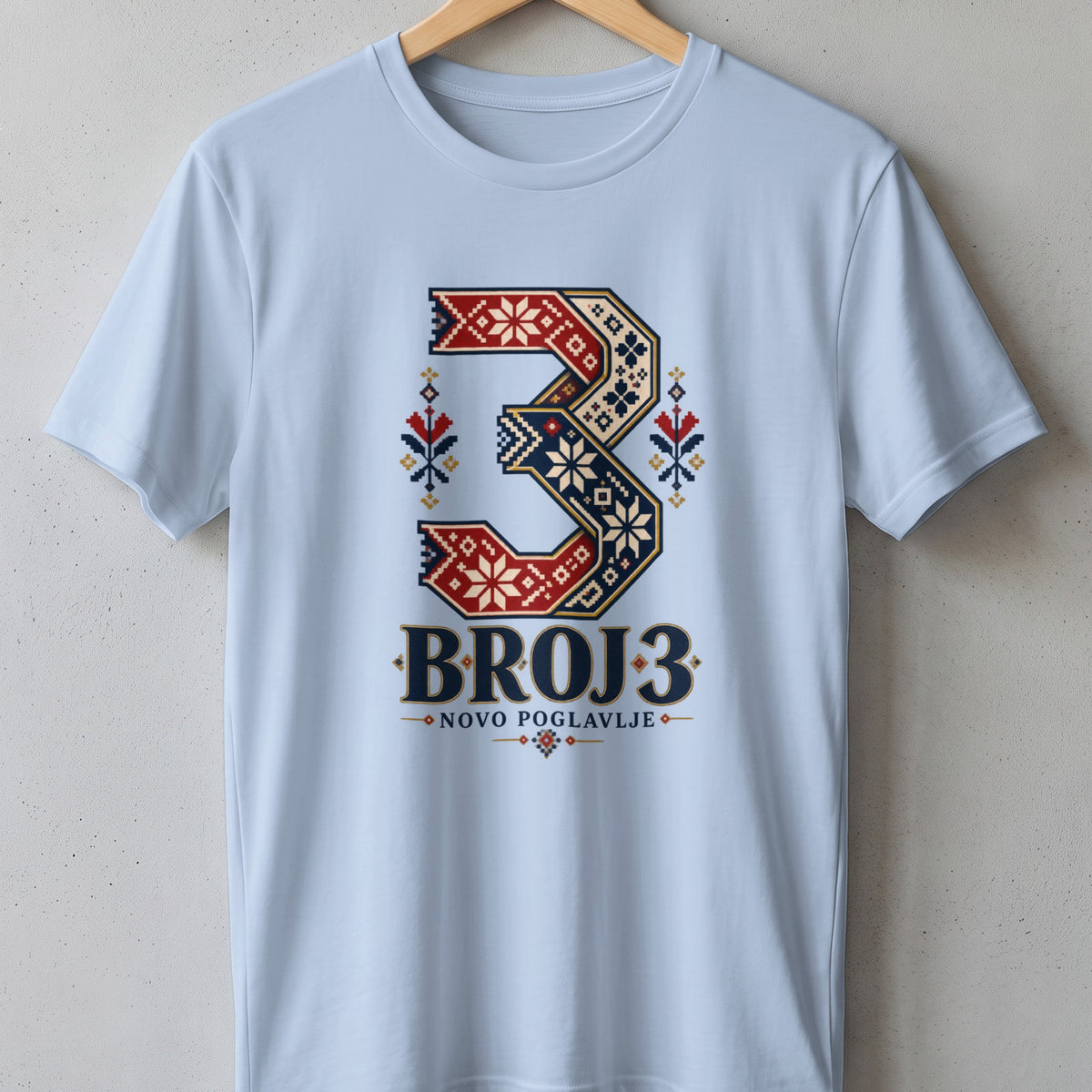 Light blue t-shirt hanging on a textured wall, featuring a large number 3 graphic with traditional folk embroidery patterns.
