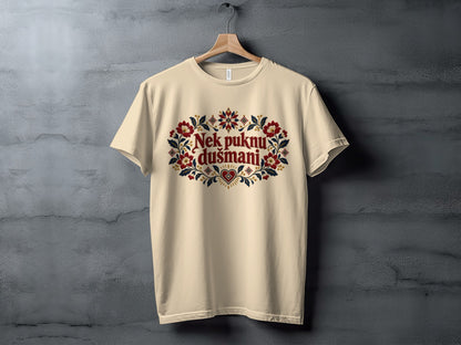 Beige t-shirt hanging on a wooden hanger against a gray textured wall, featuring red and blue folkloric embroidery graphics and text that reads Nek puknu dušmani.