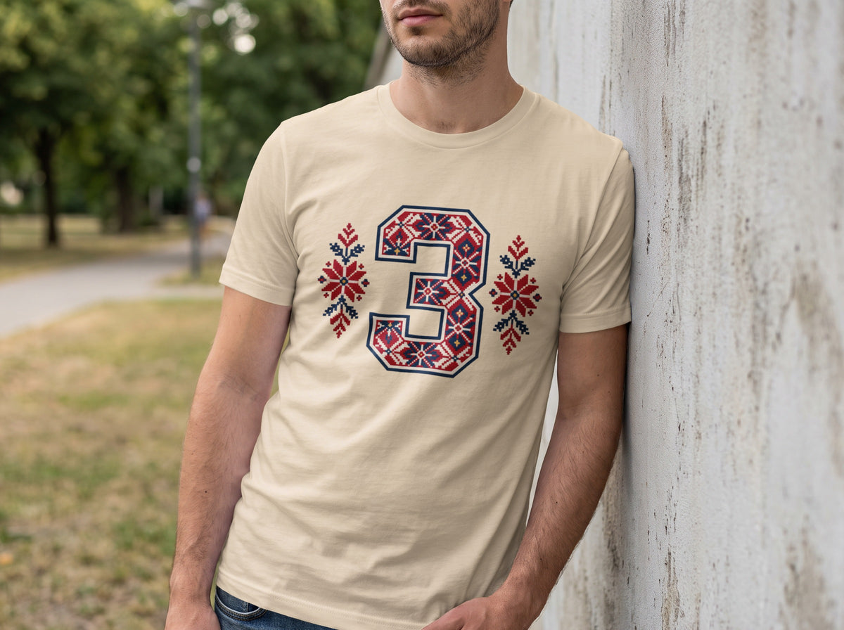 A man wearing a cream t-shirt featuring a large number three in red, white, and blue traditional folk embroidery patterns.