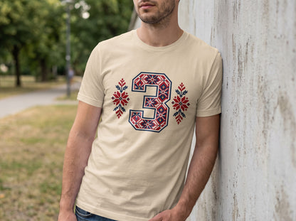 A man wearing a cream t-shirt featuring a large number three in red, white, and blue traditional folk embroidery patterns.