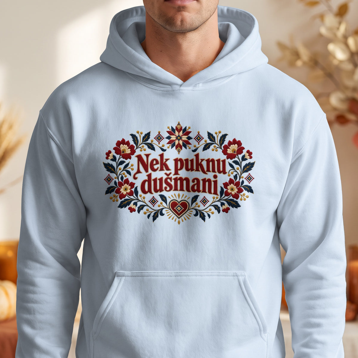 A person wearing a light blue hooded sweatshirt featuring an embroidered floral wreath and the text Nek puknu dušmani in red.