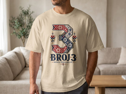 A man wearing a beige t-shirt featuring a stylized number three with red, blue, and white folk embroidery patterns and the text BROJ 3 NOVO POGLAVLJE.