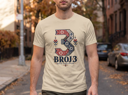 A man wearing a cream t-shirt featuring a stylized number three with folk patterns and the text BROJ 3.