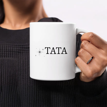 White mug with "TATA" and sparkles held by a person wearing a black sweater.