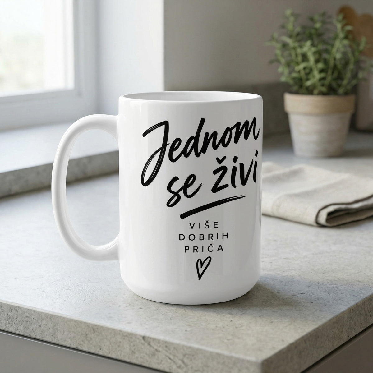 White mug with black script text "Jednom se živi" and "Više dobrih priča" with heart.