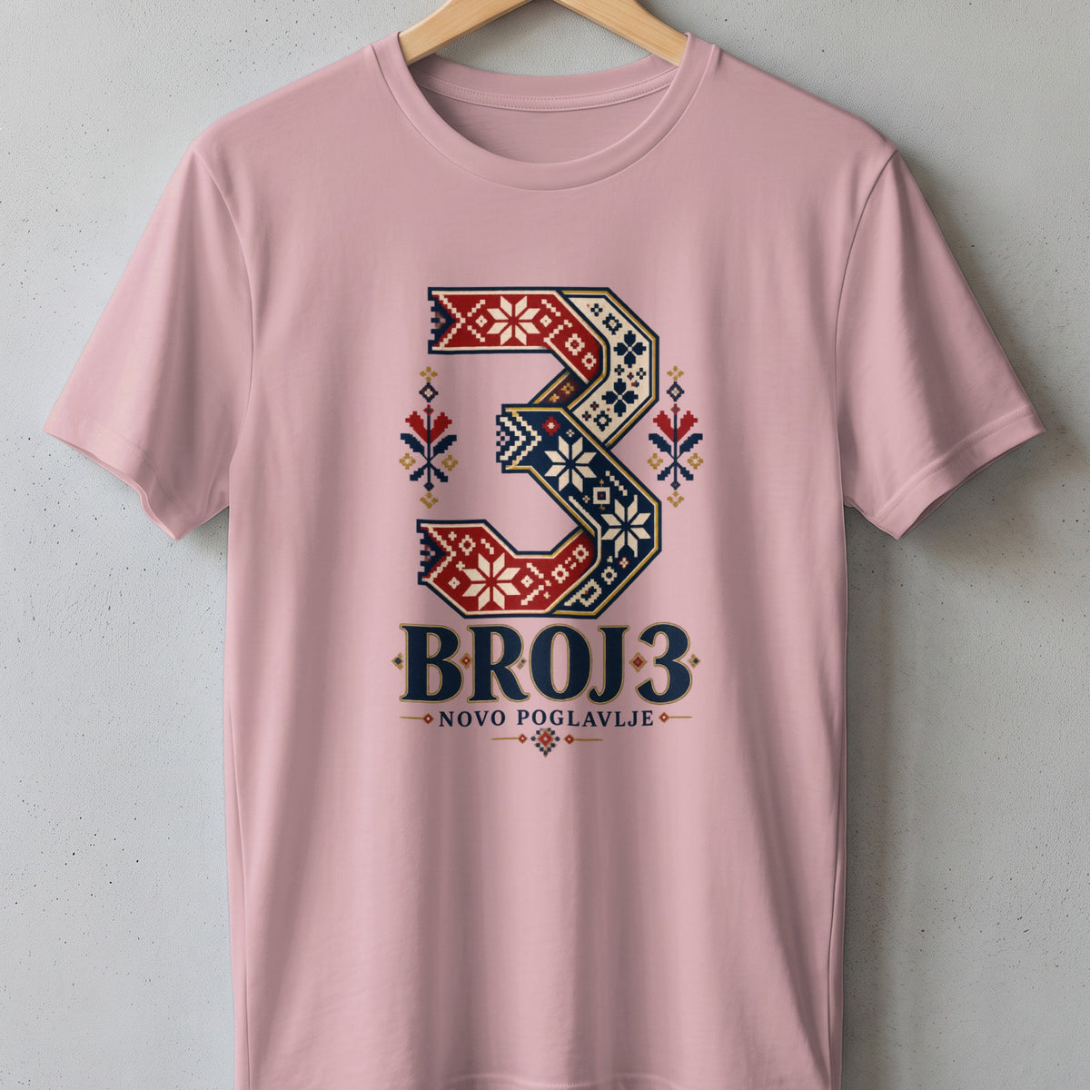 Pink short-sleeve t-shirt on a wooden hanger, featuring a large number three with traditional geometric embroidery patterns and the text BROJ 3 NOVO POGLAVLJE.